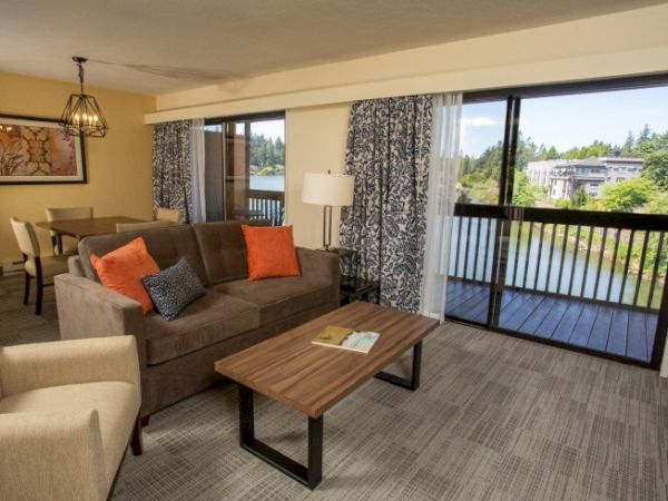 A cozy living room features a brown sofa, colorful cushions, a wooden table, a lamp, and a balcony with a scenic view outside.