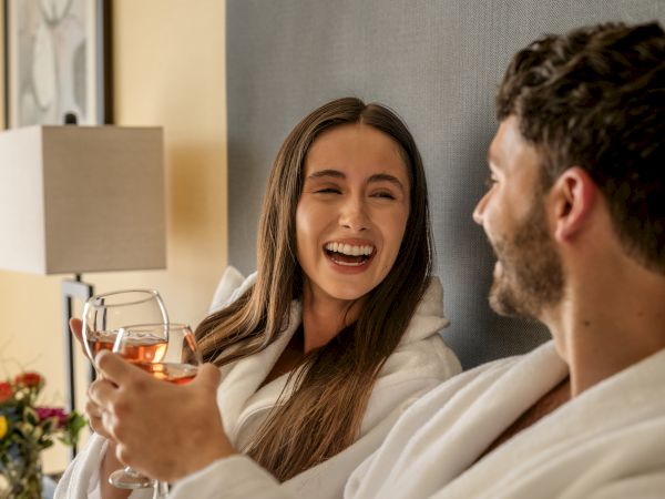 A couple in bathrobes enjoys wine, smiling and relaxing in a cozy room setting with a lamp and flowers in the background.