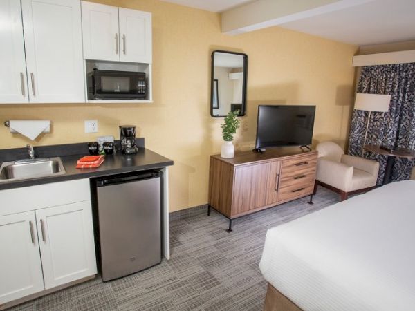 The image shows a hotel room with a kitchenette, featuring a sink, mini-fridge, microwave, TV, and bed. The decor is modern and neutral.
