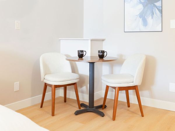 A small breakfast nook with a round table and two white upholstered chairs on a light wood floor, mugs on the table, and a framed artwork on the wall.