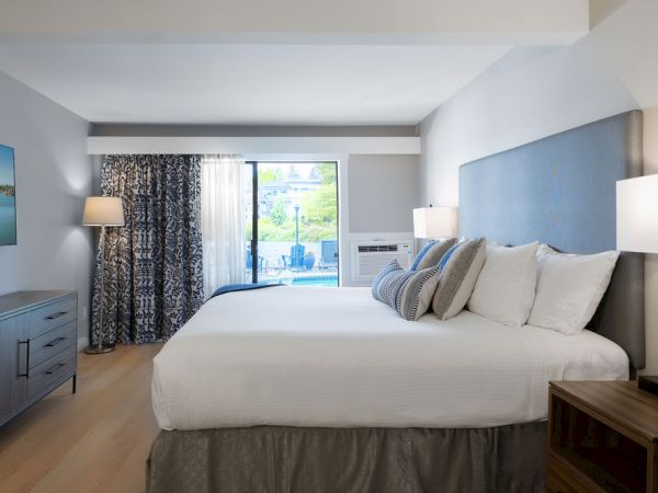 A bright hotel room with a large bed, gray headboard, two nightstands with lamps, a dresser, a wall-mounted TV, and a balcony seen through sliding doors.