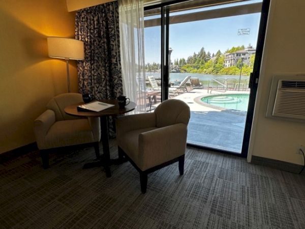 The image shows a room with two chairs, a table, a lamp, and a view of a pool and outdoor scenery through a large window or door.