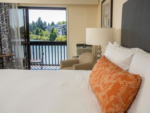 A cozy bedroom with a large bed, orange pillow, lamp, and balcony overlooking a scenic view of trees and water outside.
