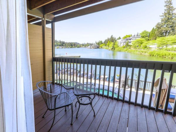 A balcony with a small round table and two chairs overlooking a lake and green shoreline under a sunny sky. Top it at 140 characters, ending the sentence.