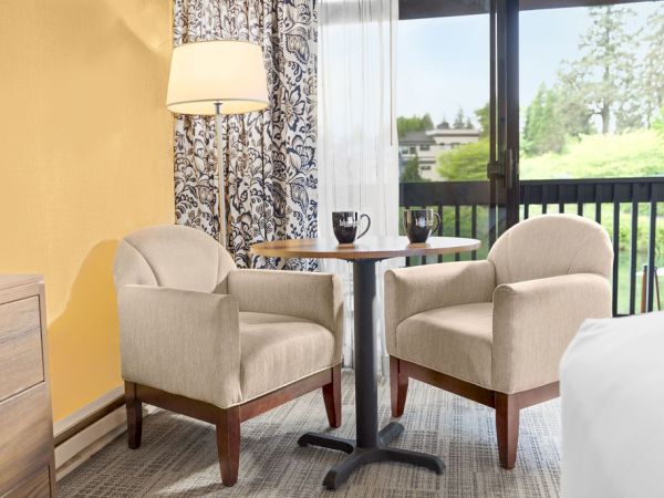 Two beige armchairs and a small round table with two cups sit by a window/door in a bright hotel room, a dresser to the left, and a yellow wall.
