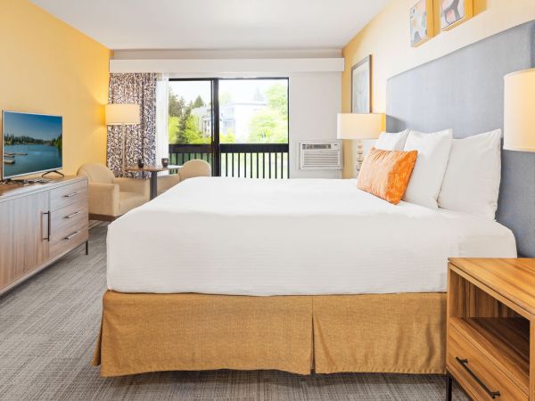 A bright hotel room with a large bed, yellow and gray accents, two nightstands, a desk, a flat-screen TV, and a balcony view.