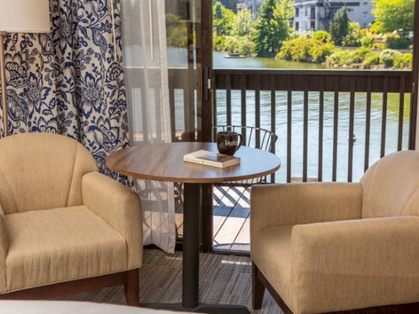 Two beige chairs face a round table with a book and mug on it, near a window overlooking a scenic lake view.