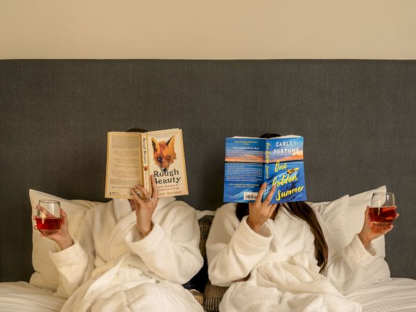 Two people in robes are lying on a bed, reading books, each holding a drink, with their feet up, covering their faces with the books.