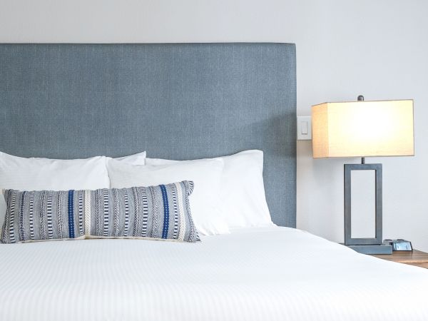 A tidy hotel-style bedroom with a white bed, blue-gray headboard, decorative throw pillow, and a bedside lamp on a wooden nightstand.