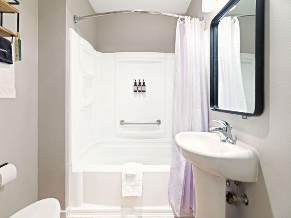 A small bathroom with a white shower/tub combo, a curved shower curtain, a pedestal sink, a mirror, and a few toiletries on the shelf, not much clutter.