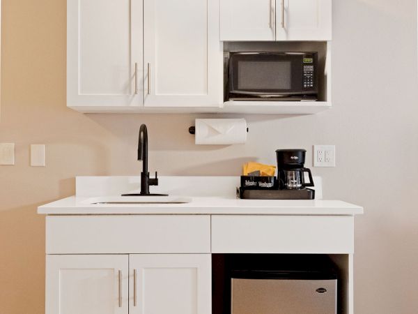 A compact kitchenette with white shaker cabinets, a small sink, a microwave, a coffee station, and a mini fridge beneath a clean countertop.