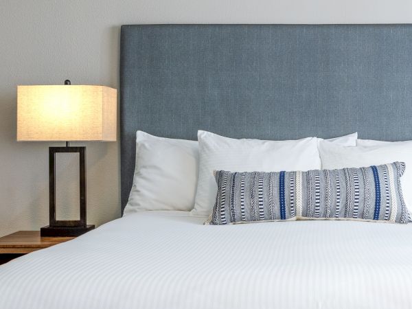 A neat hotel-style bedroom with a large blue upholstered headboard, white bedding, a patterned lumbar pillow, and a wooden nightstand with a modern lamp.