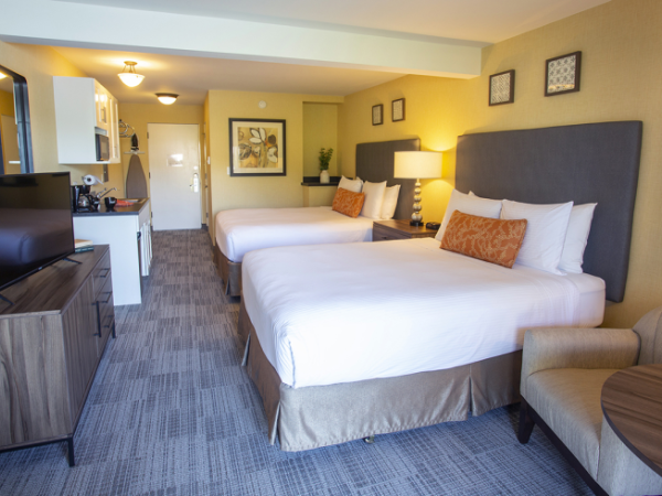 The image shows a hotel room with two beds, a TV, a small kitchenette, and a seating area, decorated warmly with modern design elements.