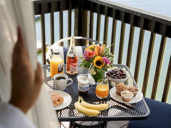 A balcony breakfast setup with orange juice, coffee, pastries, fruit, and flowers. A hand is pulling back a curtain to reveal the scene.