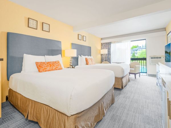 Two double beds in a bright hotel room with yellow walls, blue headboards, white linens, orange accent pillows, a desk, and a balcony view.