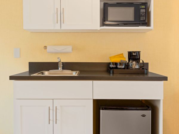 A compact kitchenette with white cabinets, a black countertop, a sink, a microwave in upper cabinet, a small coffee area, and a mini fridge beneath; yellow wall behind.