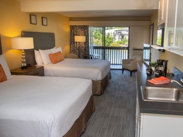 A hotel room with two beds, a kitchenette, and a balcony view of a building and trees. Bright decor with pillows adds a cozy touch.