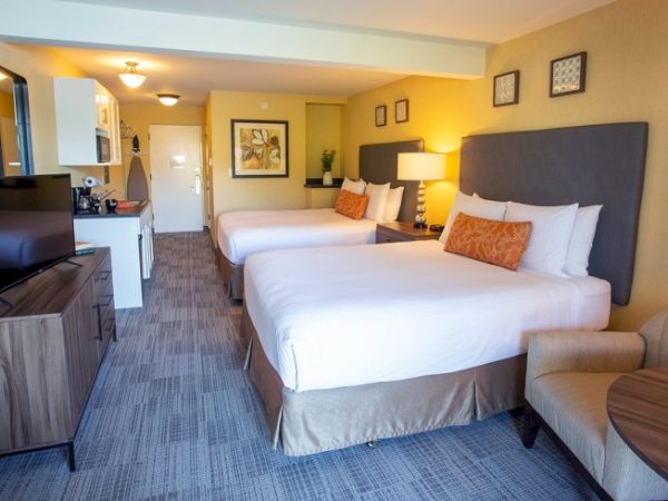 A hotel room with two double beds, a TV, a kitchenette, and comfortable seating, decorated in warm tones with modern furnishings.