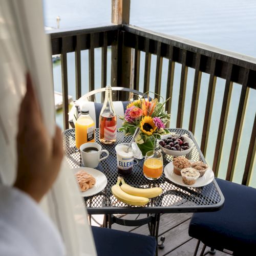 A breakfast spread on a patio table includes fruit, muffins, juice, and coffee, with a lake view and a hand pulling a curtain aside.