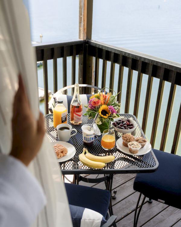 A breakfast spread on a patio table includes fruit, muffins, juice, and coffee, with a lake view and a hand pulling a curtain aside.