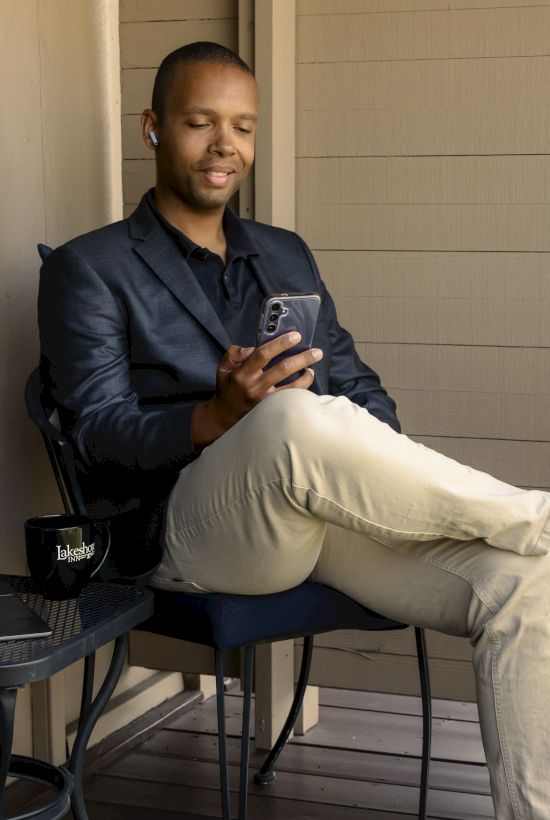A person is sitting on a chair, looking at a phone. There's a table with a laptop and mug nearby on a balcony or patio.