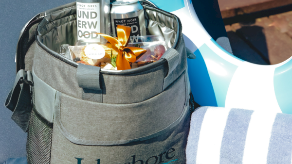 A Lakeshore Inn cooler bag with wine bottles, snacks, and a gift bow, next to pool towels and a blue slide.