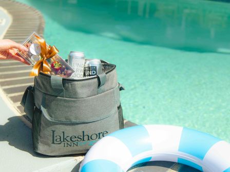 A Lakeshore Inn cooler bag by a pool holds drinks and snacks, with a hand placing items inside, and a blue and white pool float nearby.