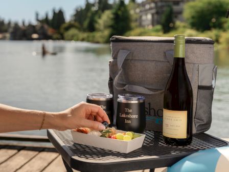 Picnic setup by a lake, featuring a cooler bag, wine bottle, snack tray, and two cups on a table.