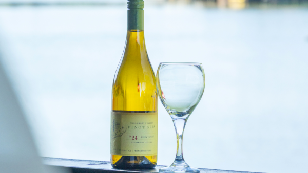 A bottle of white wine with a stemmed glass on a patio table near water, with boats blurred in the background.