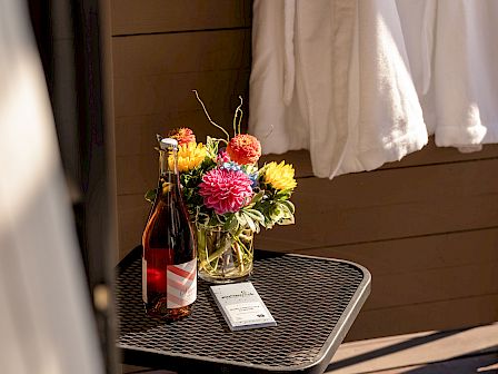 A small table holds a flower arrangement, a wine bottle, and a white paper, next to a white curtain or fabric.