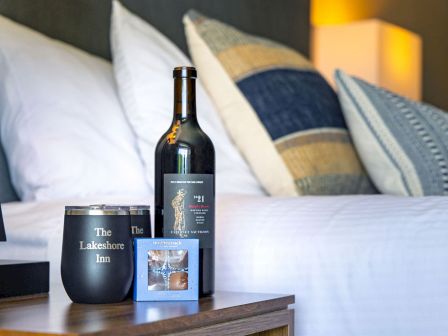 A cozy bedside setup with a bottle of wine, a glass, a candle-like lamp, and framed photos on a wooden nightstand near a bed.