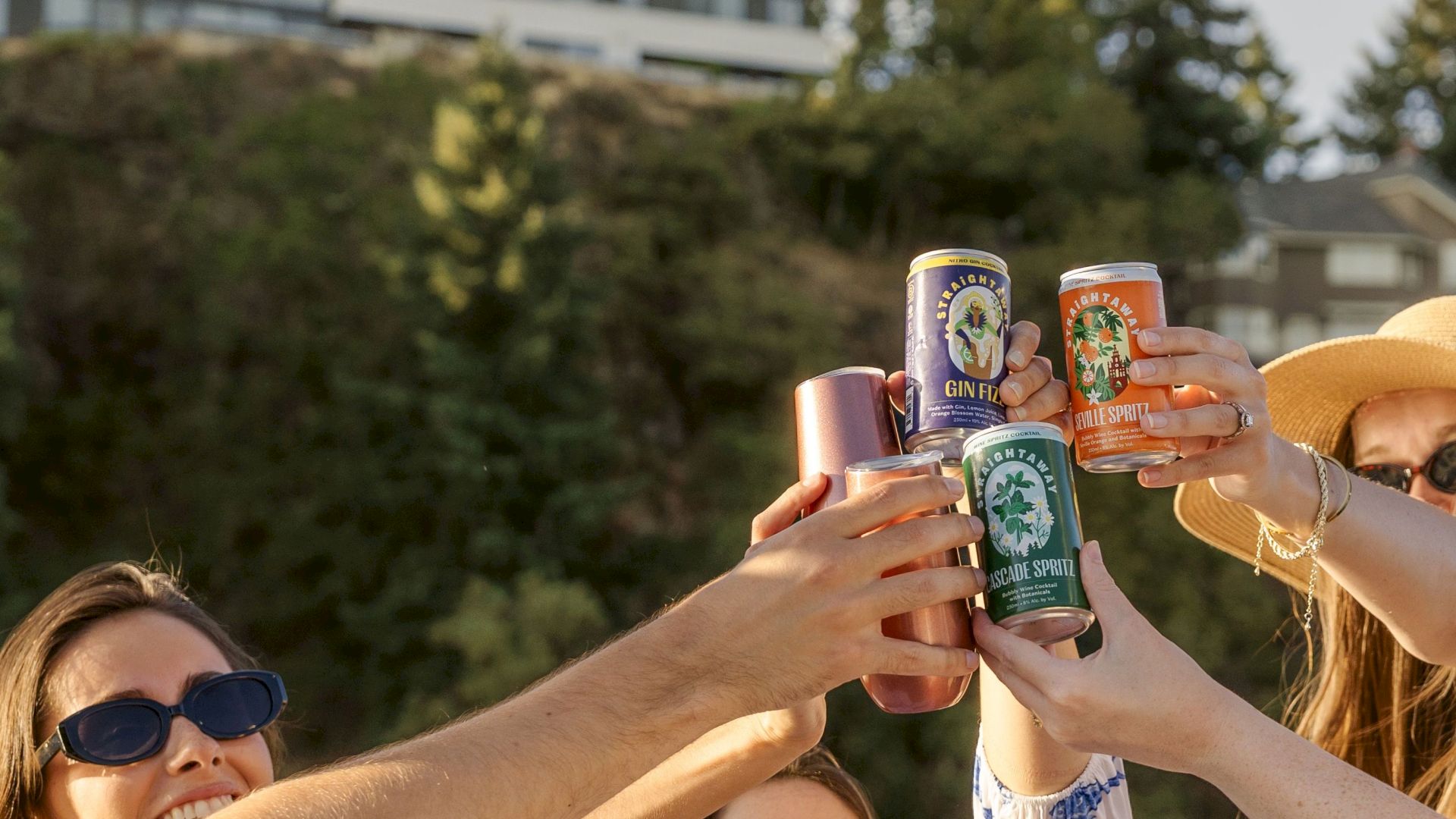 A group of people outdoors raising cans in a toast, with trees and buildings in the background, under a sunny sky.