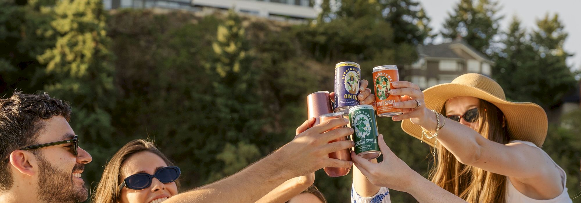 A group of people outdoors raising cans in a toast, with trees and buildings in the background, under a sunny sky.