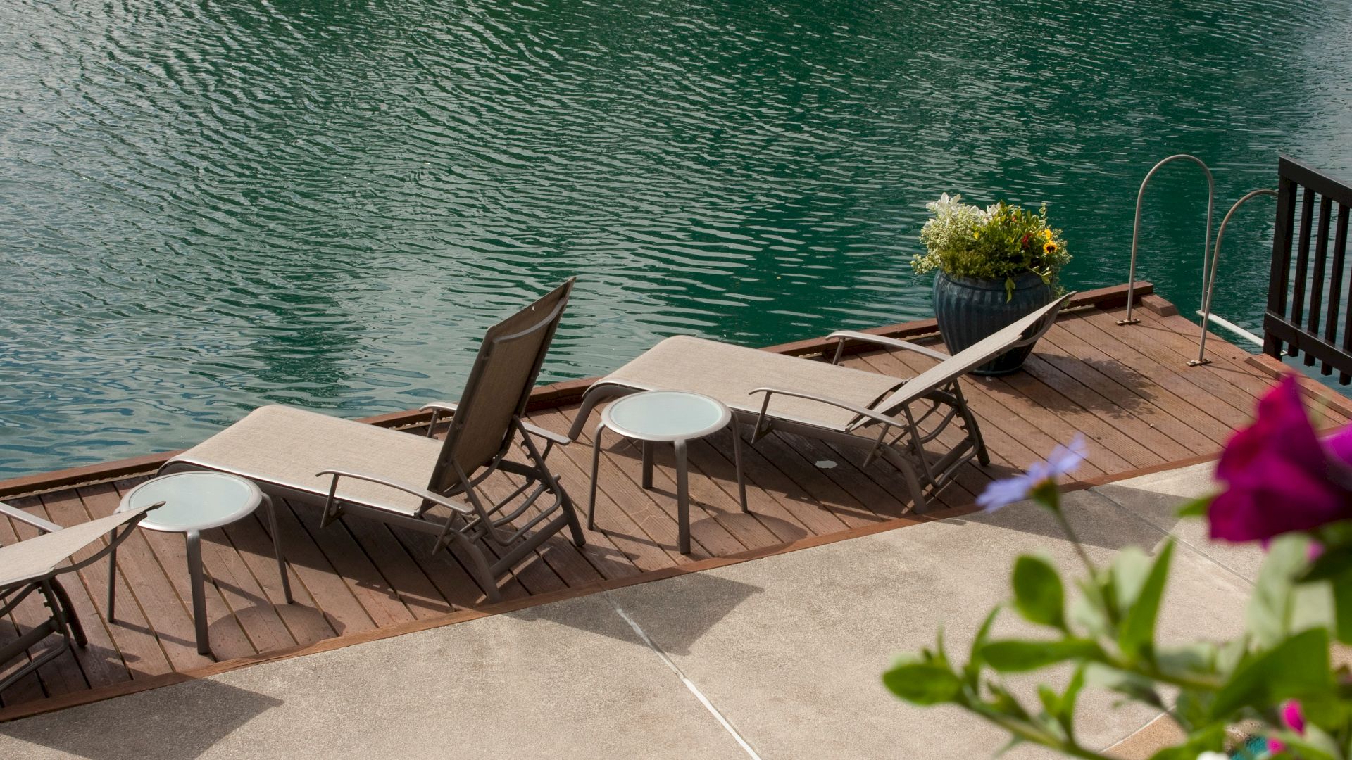 Lakeside scene with lounge chairs, small tables, and a potted plant on a wooden deck by the water, with flowers in the foreground.