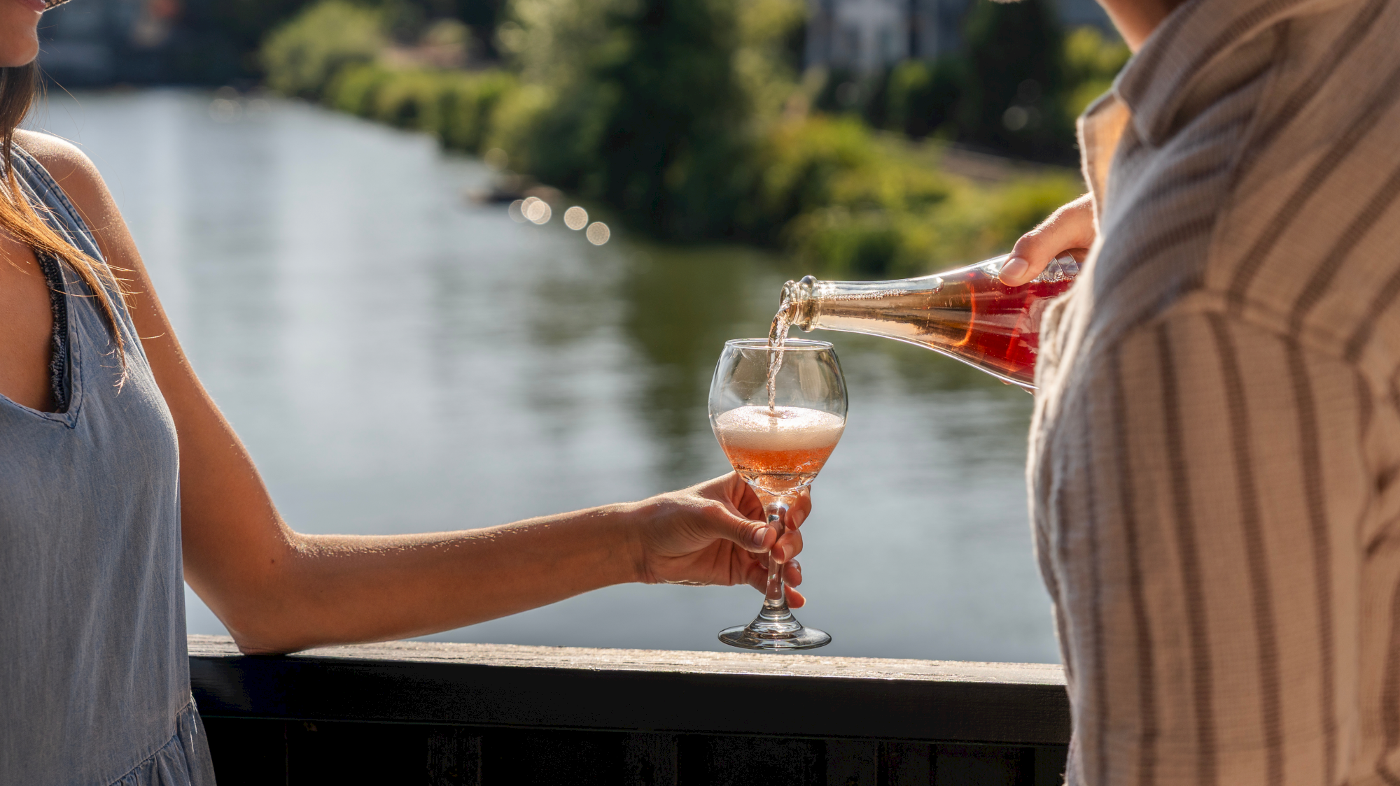 Two people enjoying a drink by a river, with one pouring a beverage into a glass held by the other.