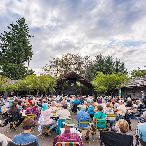 A crowd of people is seated outdoors, facing a stage in a park or garden setting under a cloudy sky, surrounded by trees and a building.