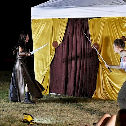 Two people are performing a sword fight in front of a tent with yellow and brown curtains, while others watch from the grass.