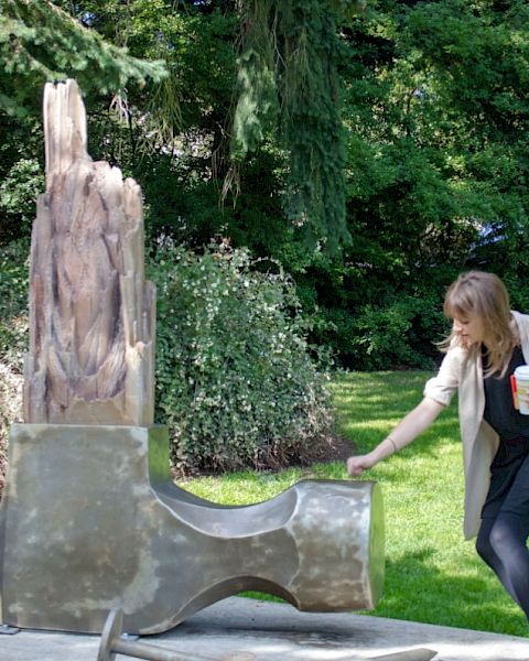 A person interacts with a modern sculpture in an outdoor park setting, holding a drink and a bag.