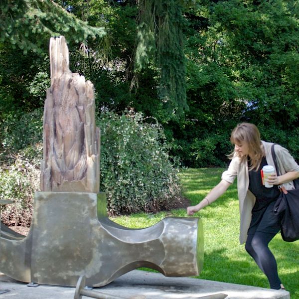 A person interacts with a modern sculpture in an outdoor park setting, holding a drink and a bag.