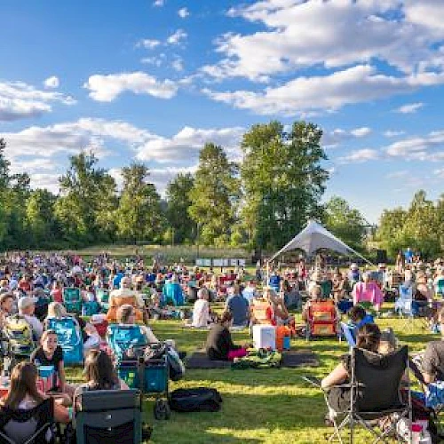 A large outdoor gathering with people seated on grass and in chairs, facing a stage under a tent, surrounded by trees and a partly cloudy sky.