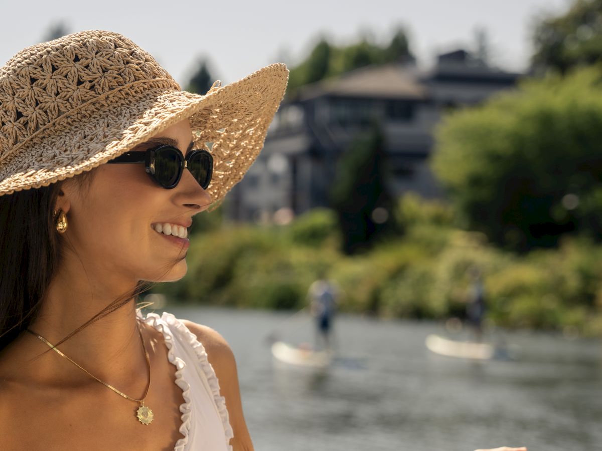 A person wearing a sunhat and sunglasses, holding a striped towel by the water, with paddleboarders in the background.