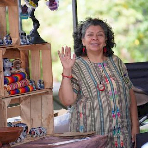 A person waving at a stall with colorful items, like pottery and textiles, displayed on wooden shelves outdoors.