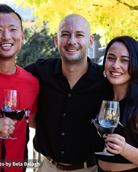 Three people smiling and holding wine glasses are posing together outdoors with autumn leaves in the background.