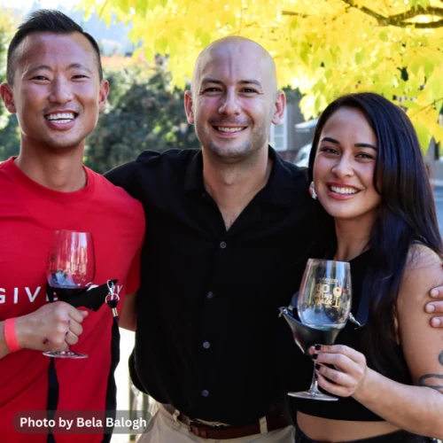 Three people smiling and holding wine glasses are posing together outdoors with autumn leaves in the background.