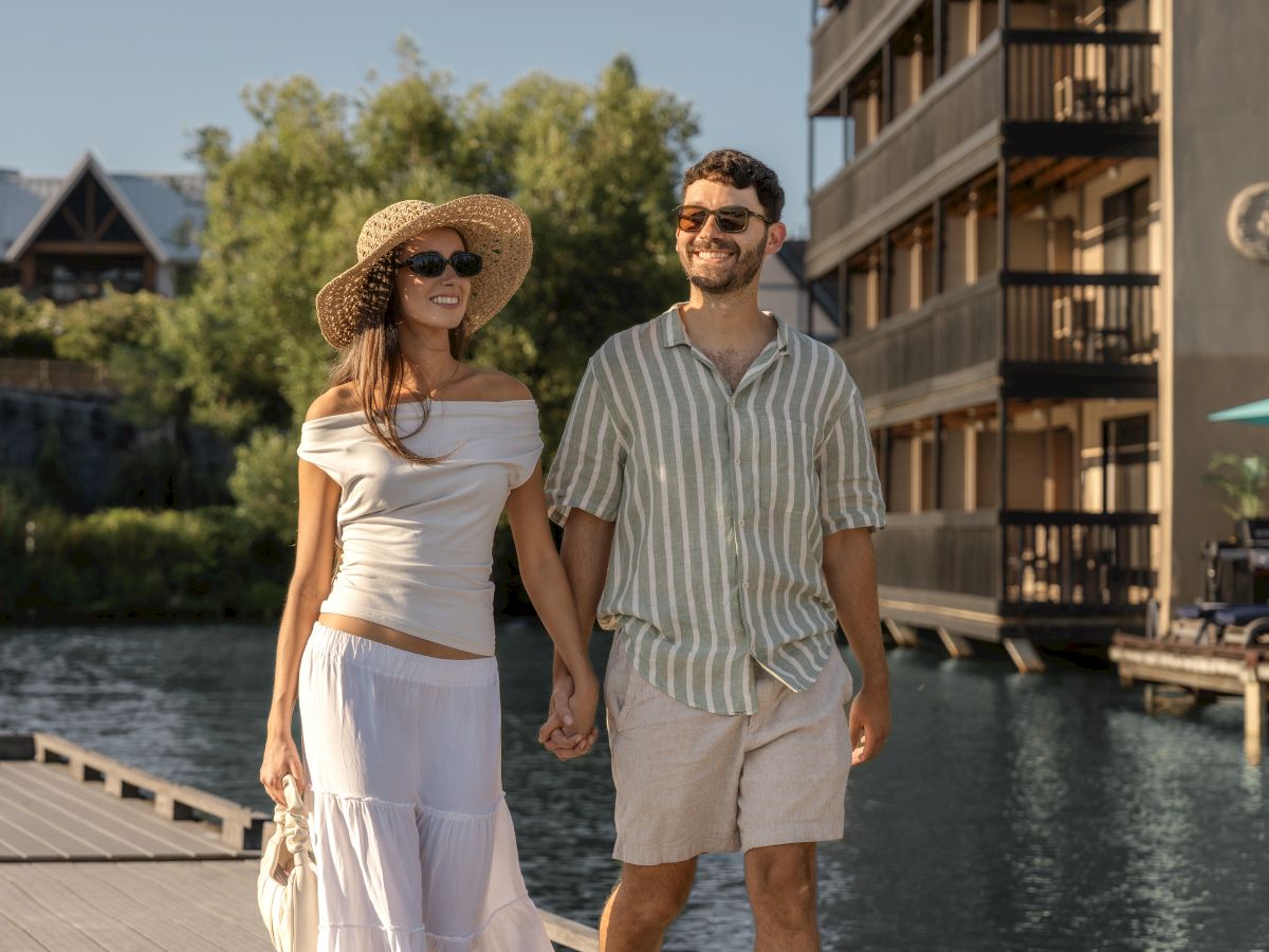 A couple holding hands, walking near water. They're dressed casually, both wearing sunglasses, and appear relaxed and happy.
