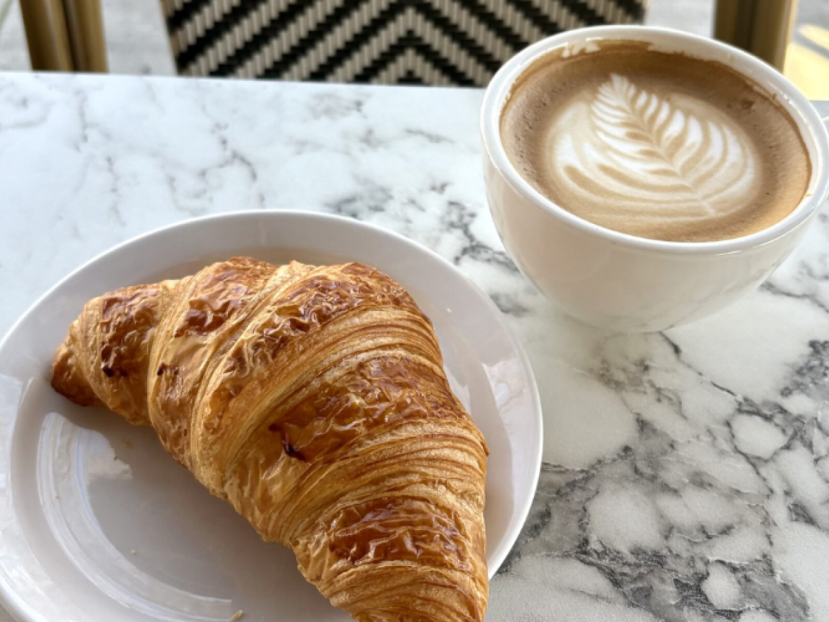 A flaky croissant on a white plate and a cup of latte with latte art sit on a marble table, providing a perfect cafe scene.