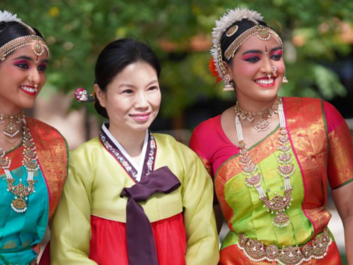 Three people are in traditional attire, two in colorful Bharatanatyam costumes and one in a hanbok, standing outdoors.