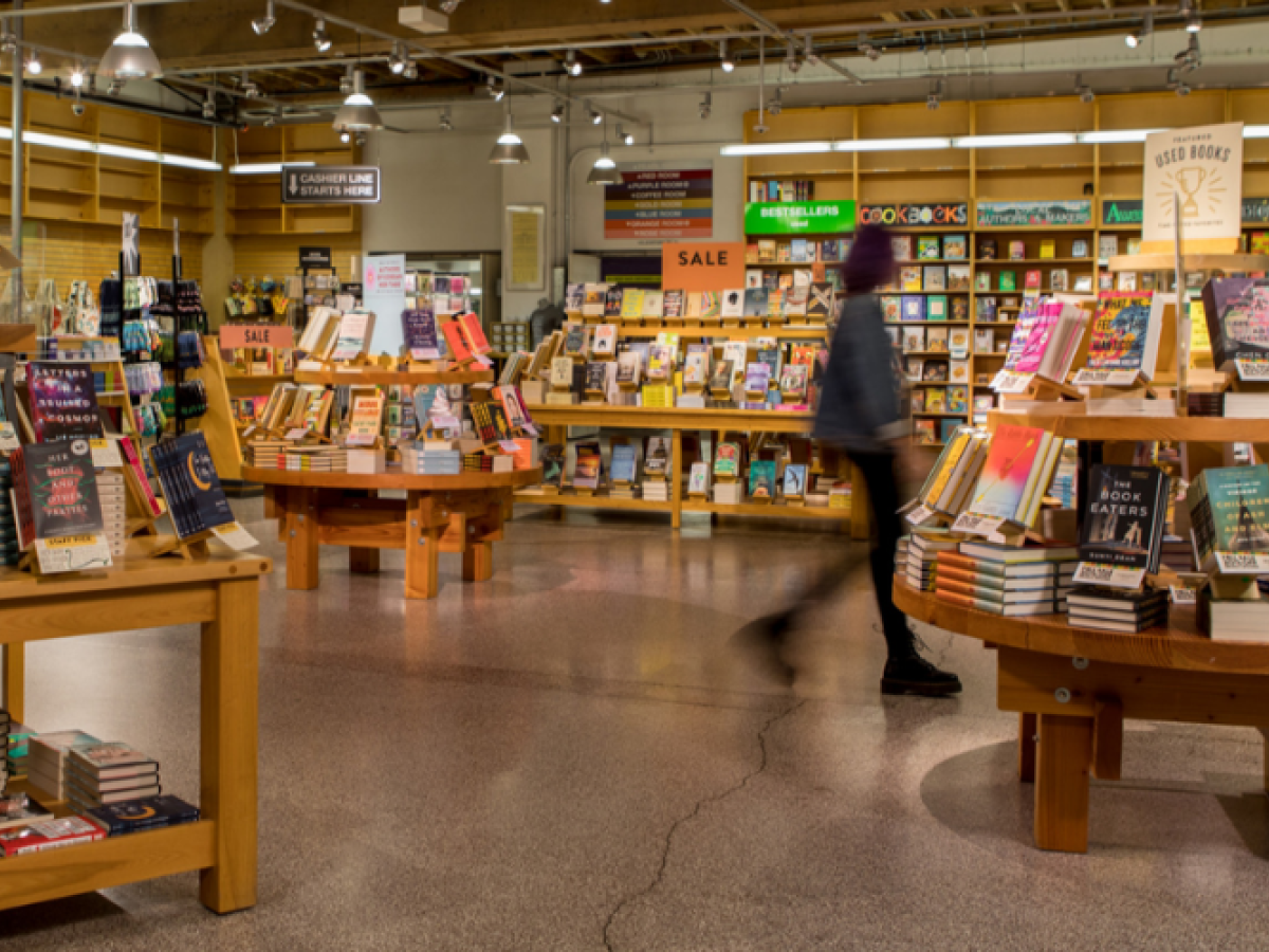 The image shows a bookstore interior with wooden shelves and tables filled with books, and a person walking by.