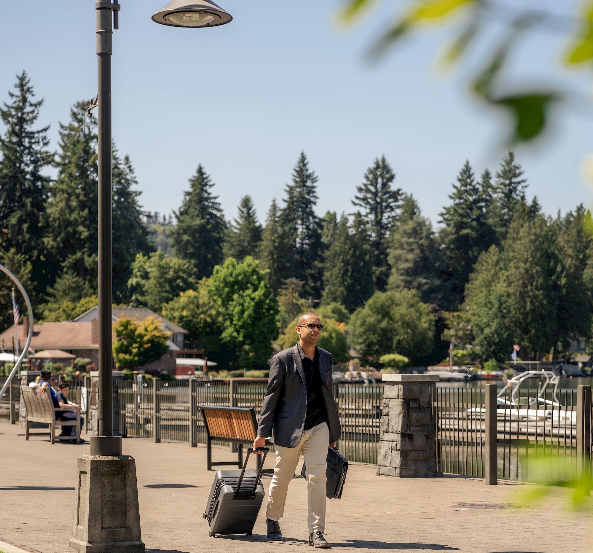 A person is walking along a riverside path with rolling luggage on a sunny day, surrounded by trees and park amenities.