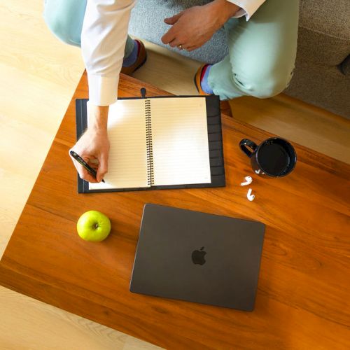 A person sits on a couch, writing in a notebook on a wooden table with a laptop, apple, coffee cup, scattered papers, and a few pills nearby.
